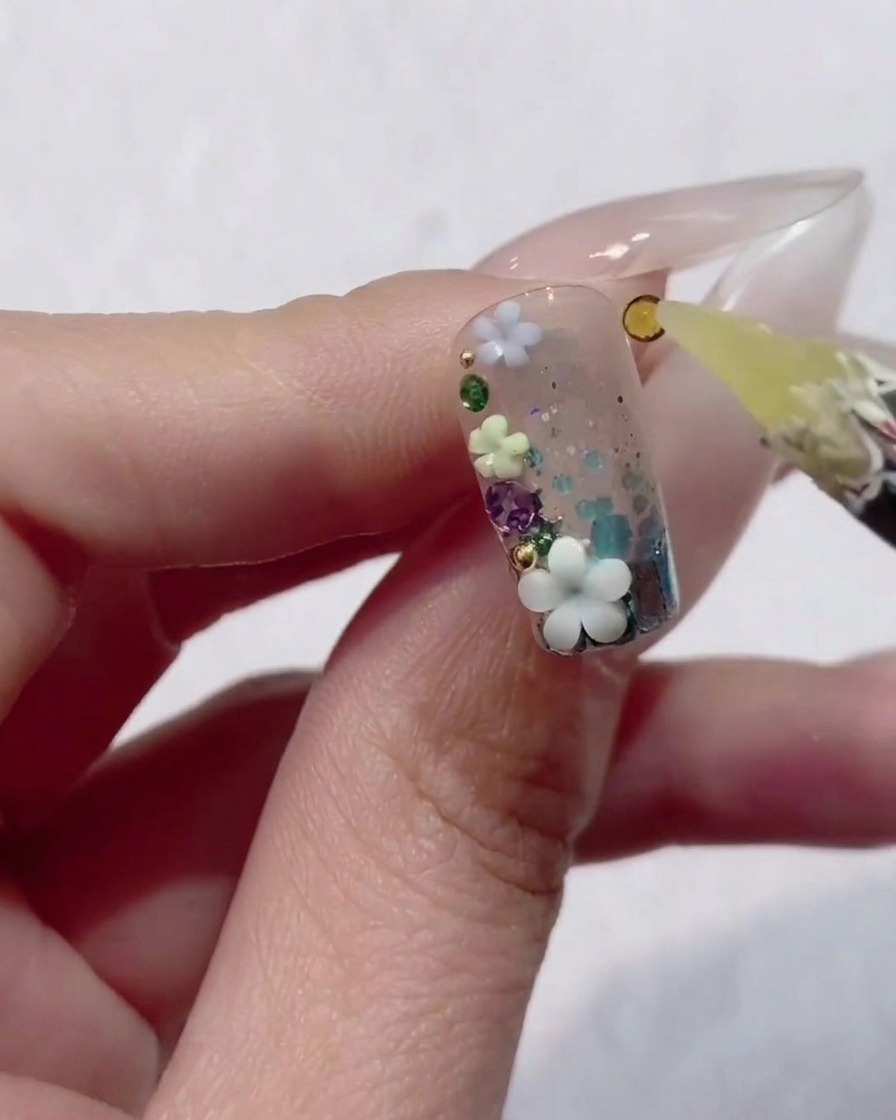 A close-up of a hand holding a fingernail decorated with clear gel, glitter, and tiny 3D flowers, while a wax tool places a small yellow rhinestone into the design.