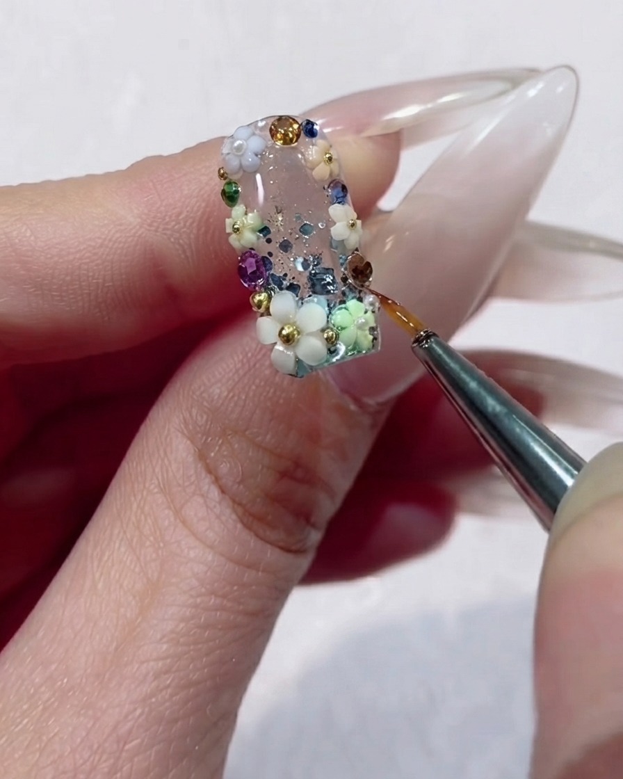 A fine liner brush precisely applying clear gel around the edges of a faceted rhinestone on a floral nail design.