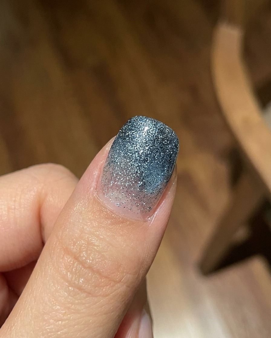 A finger displaying a translucent blue jelly gel gradient coat over a textured glittery background, shot in bright natural light.