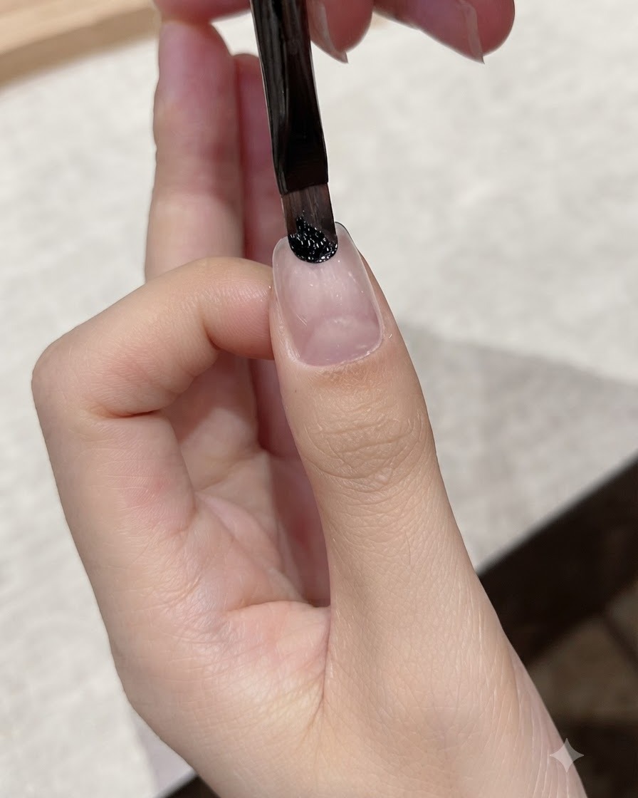 A fine liner brush applying dark black gel near the cuticle area of a natural nail.