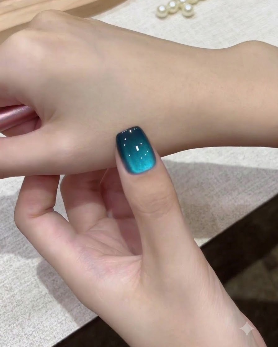 A close-up view of a single teal galaxy cat-eye nail held against the back of the hand.