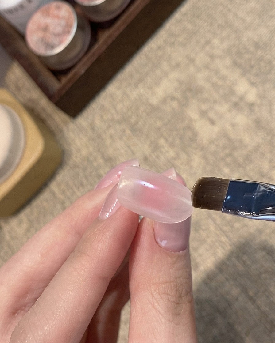 A flat brush applying a blue shimmer cat-eye gel onto the tip of a pinkish-nude nail.
