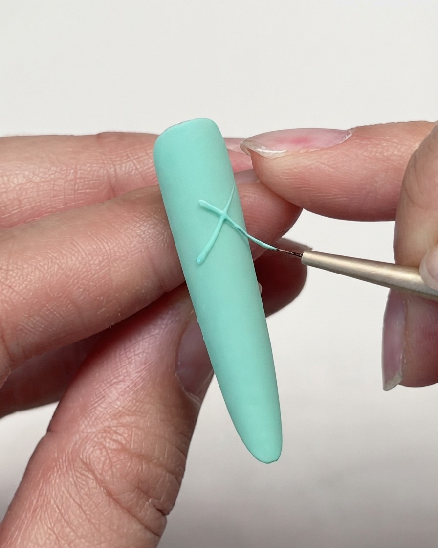 A close-up of a fine liner brush crossing gel lines on a mint green matte nail tip to start a star design.