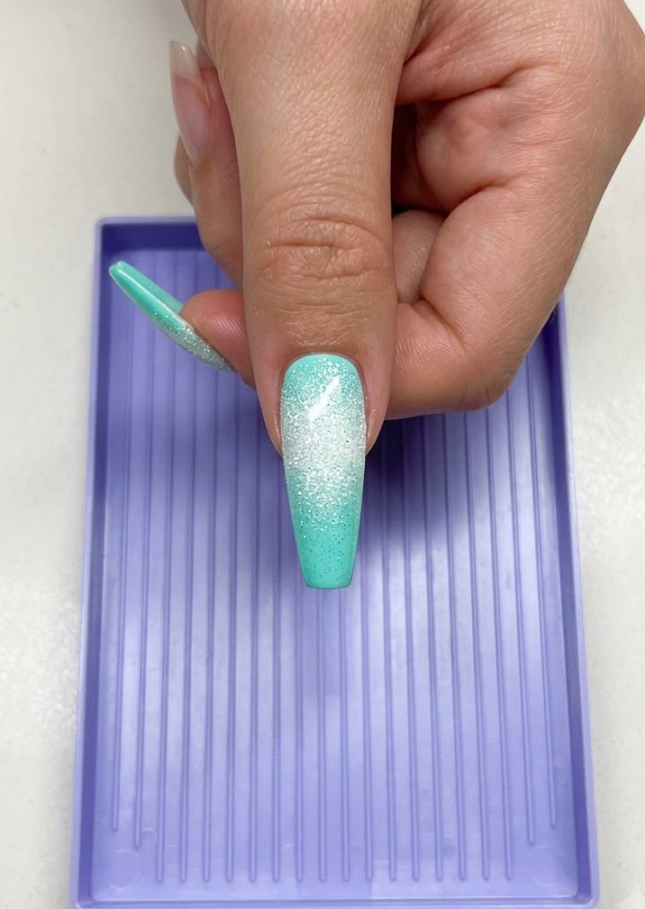 A hand holding a long mint-colored nail with a white sugary star design underneath a purple UV LED curing lamp.