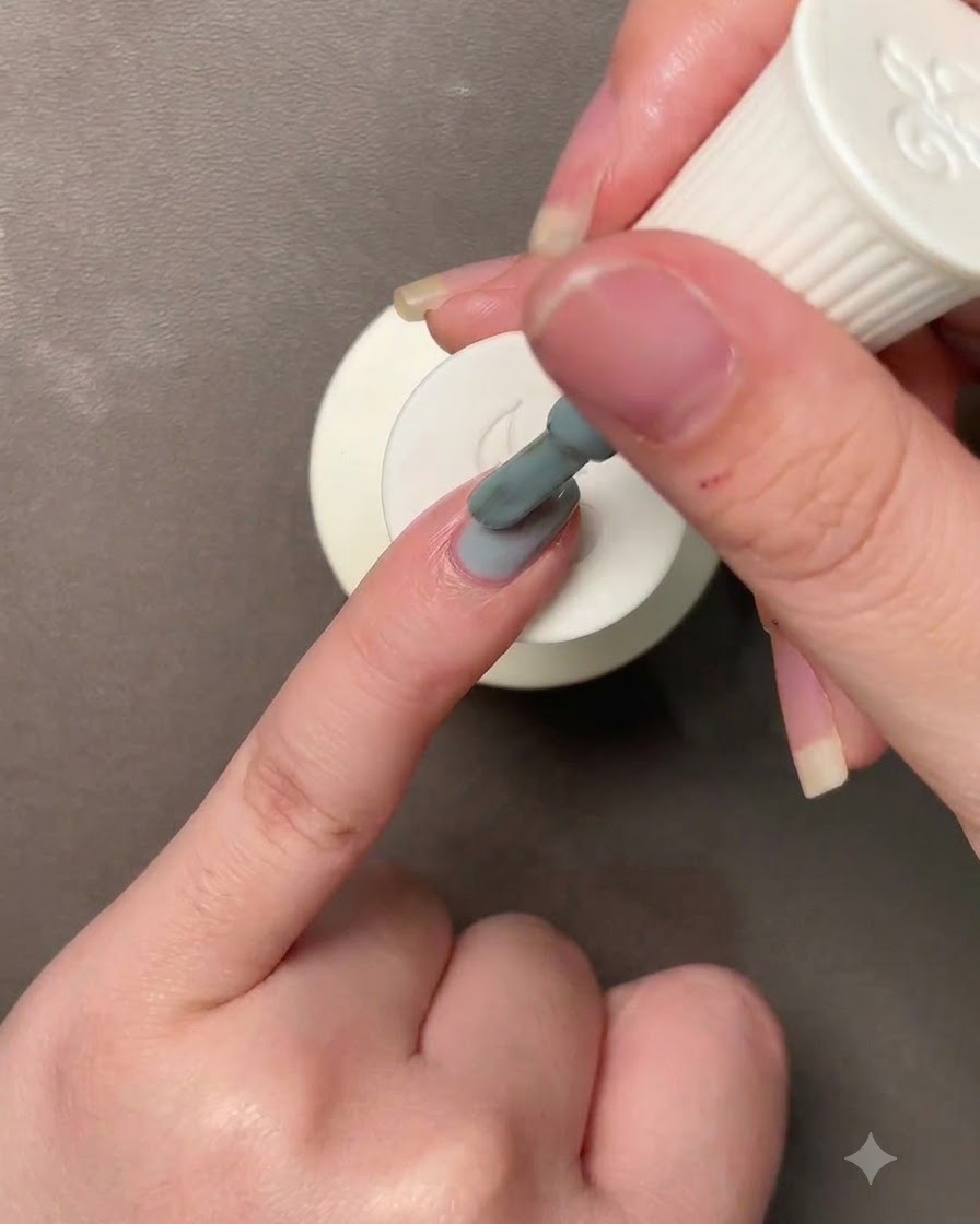 A hand holding a brush applying a grayish Morandi blue-green gel polish to a fingernail.