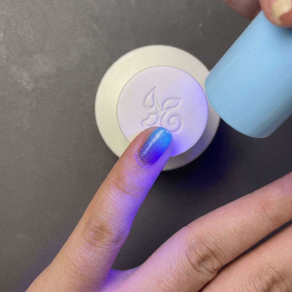 A finger with bright blue gel polish being cured under a small, handheld light blue UV/LED lamp.