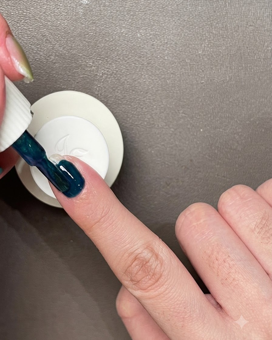 Applying a layer of dark blue-green gel polish directly onto a bare natural fingernail.