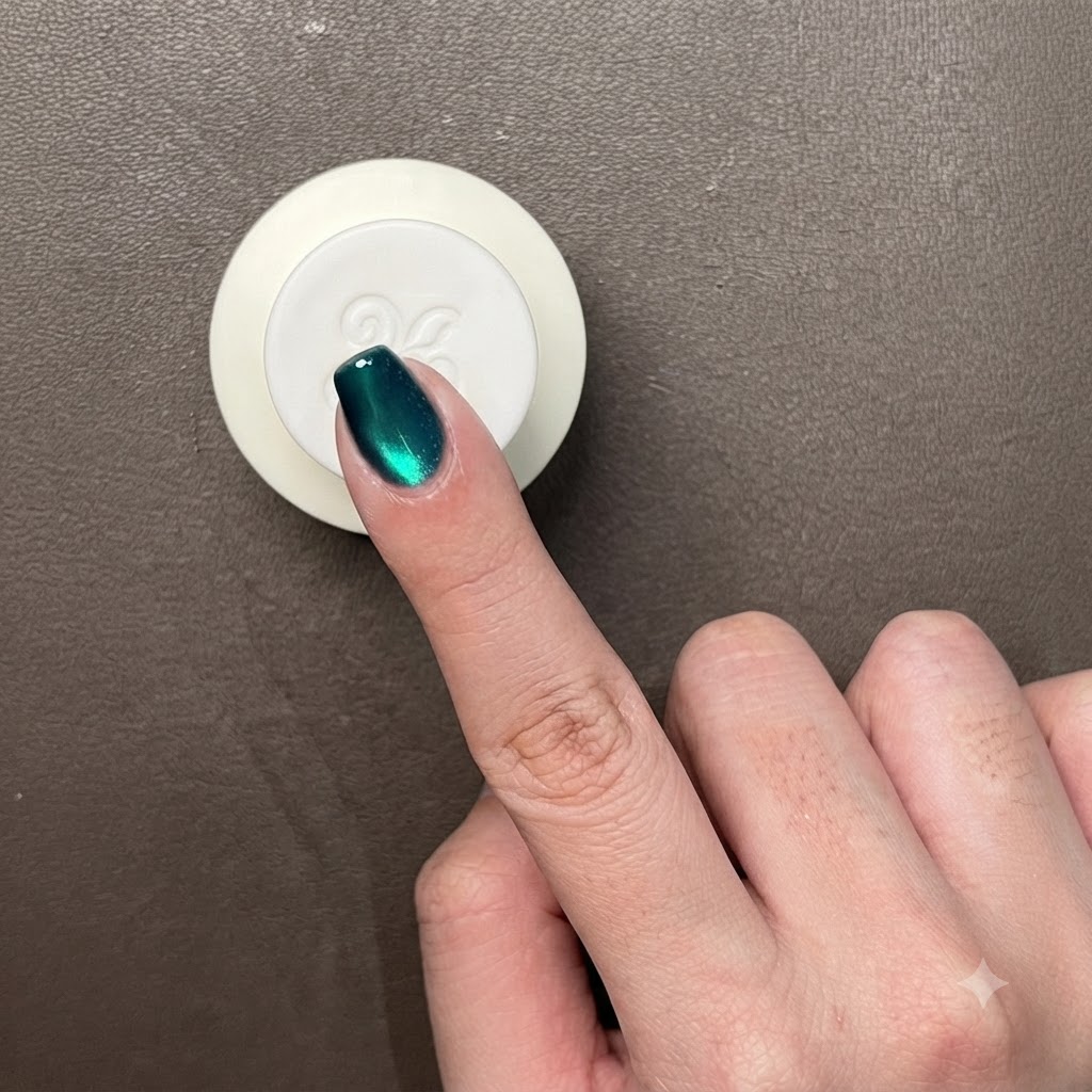 A close-up of a finger with dark metallic green cat-eye gel, showing a bright circular glass bead light reflection.