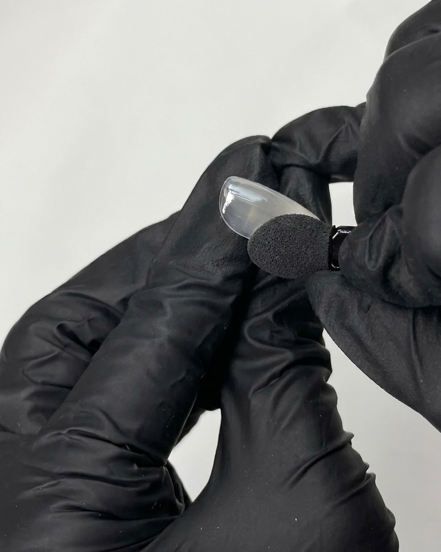 A black-gloved hand using a sponge applicator to rub iridescent chrome powder onto a smooth fake nail tip.