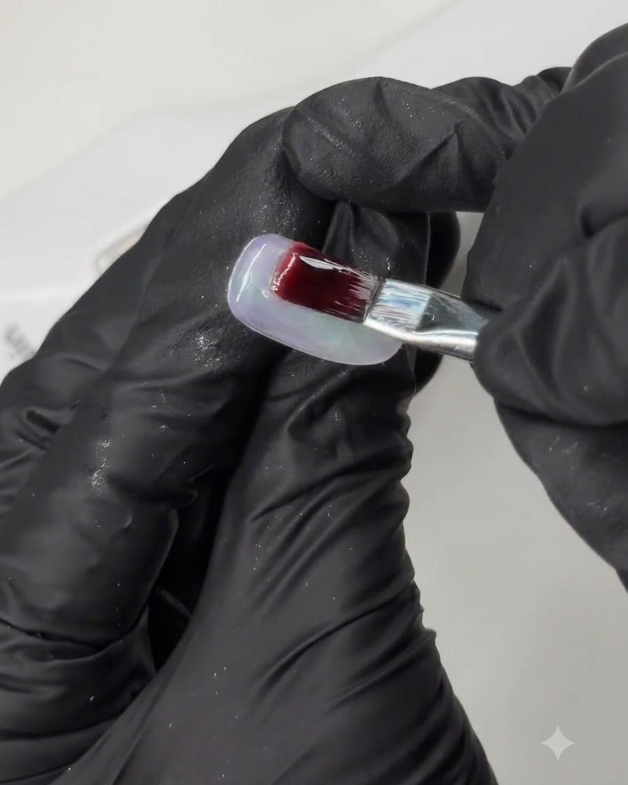 A brush applying a deep pigmented gel onto a practice nail tip during a multi-step nail art tutorial.