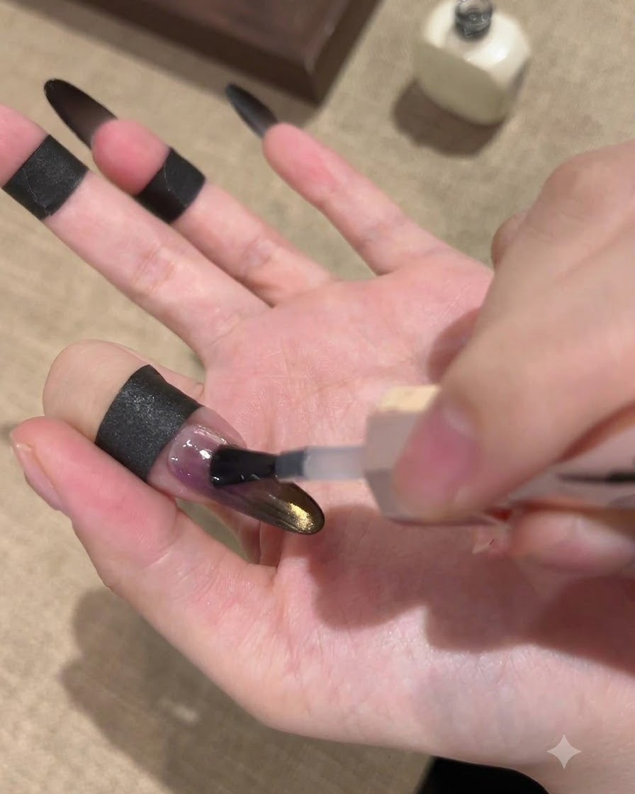 A brush applying a glossy sealing top coat over a black and gold gradient cat-eye nail.