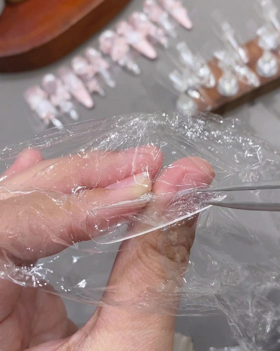 A pair of tweezers peeling away plastic wrap from a cured, textured clear nail tip.