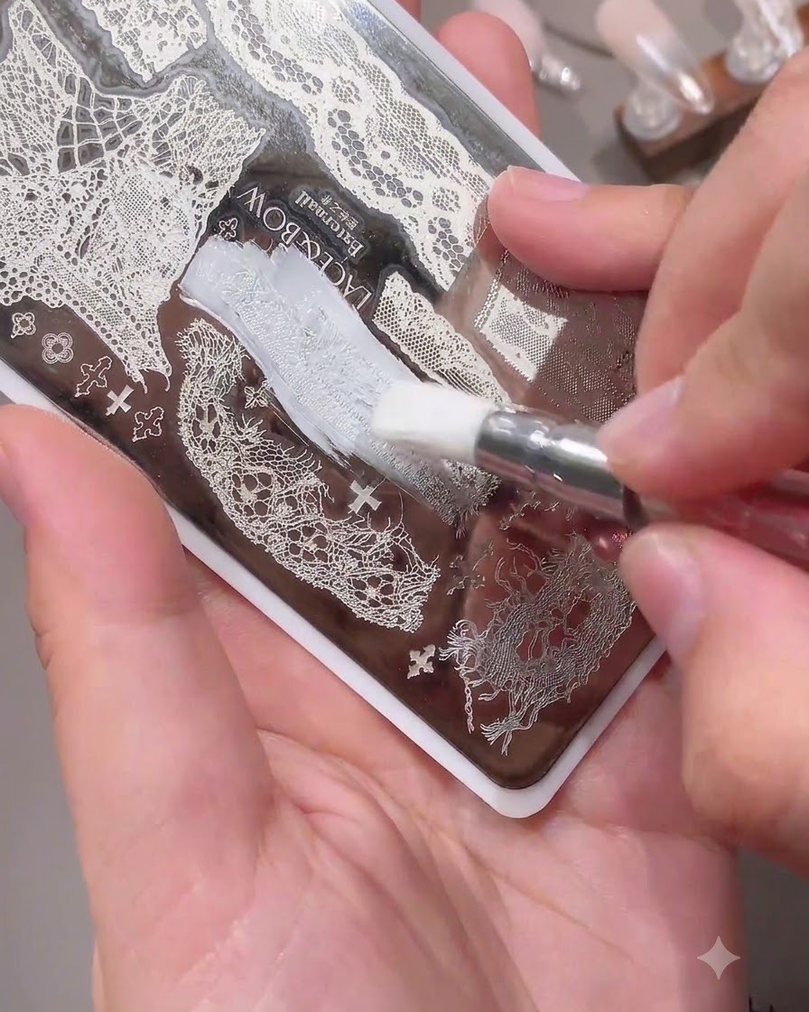 A brush applying white polish over a delicate lace pattern on a metal nail stamping plate.