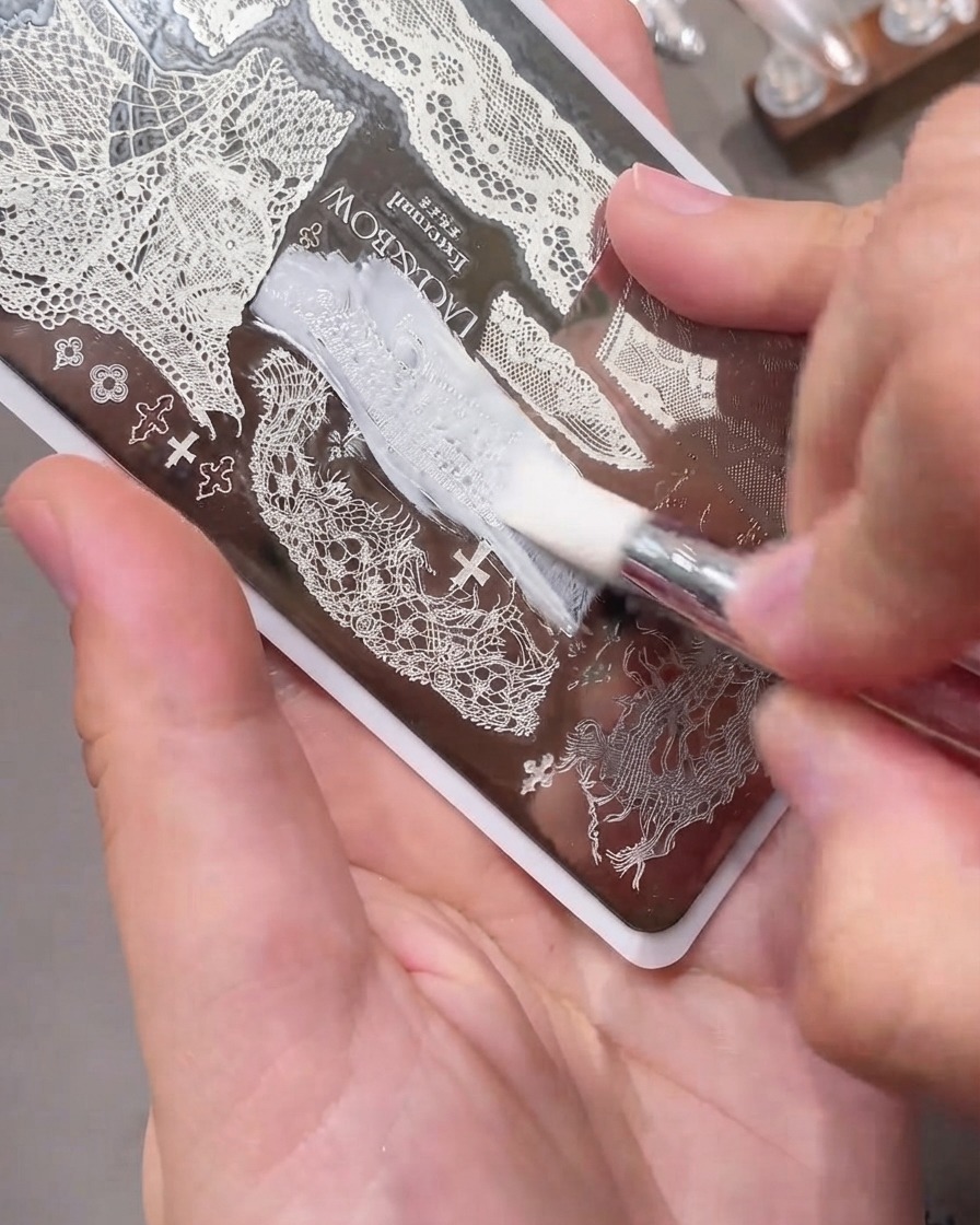 A scraper card gliding smoothly across an engraved metal nail stamping plate filled with white gel polish.