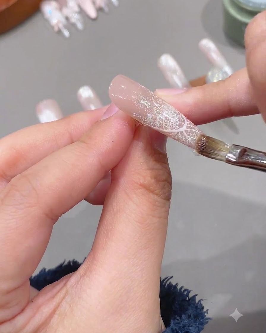 A brush applying a clear layer of soft reinforcement gel onto a prepped nail with lace design.