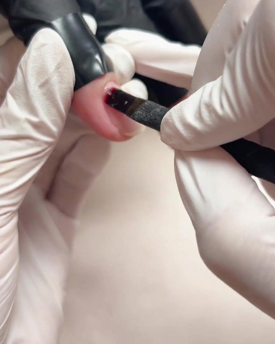 A close-up view of a gloved hand using a small brush to apply clear base gel onto a natural fingernail.