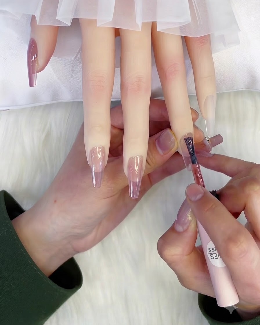 A brush applying a second coat of translucent pink gel polish onto a nail.