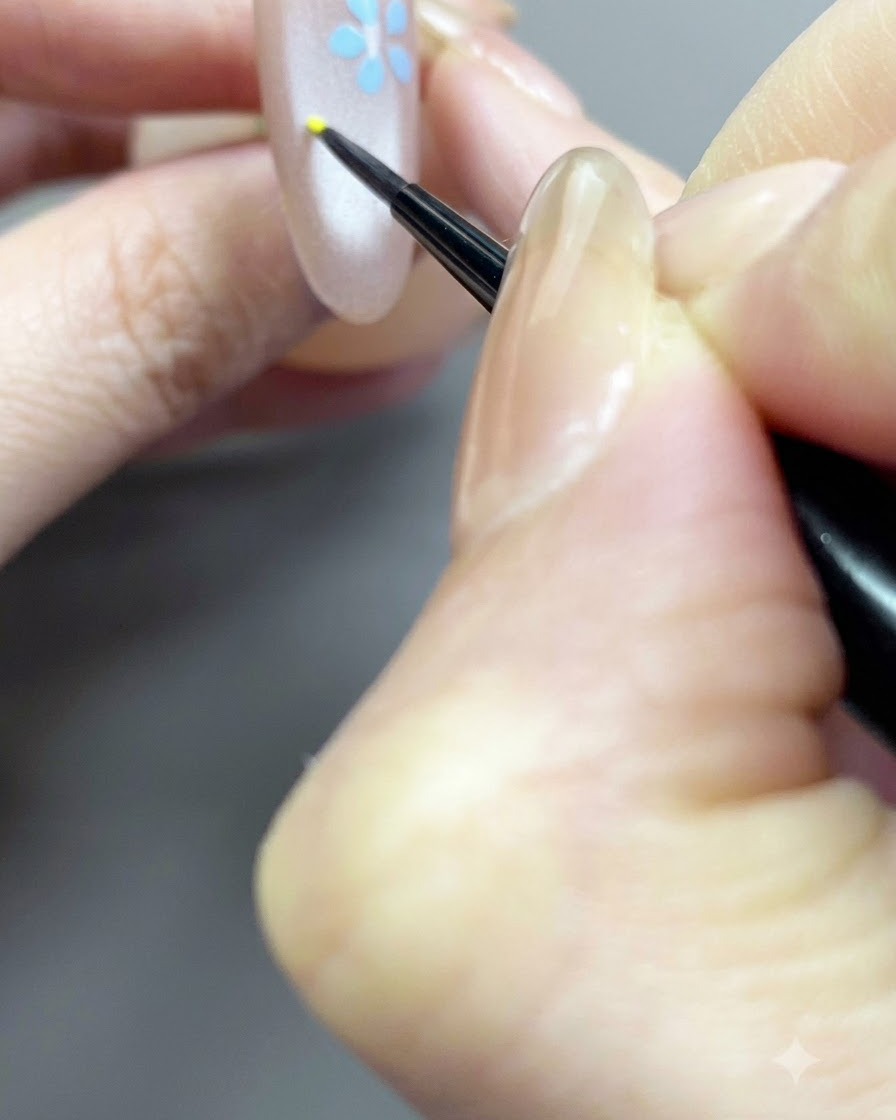 A fine brush detailing a small yellow center dot for a floral design on a nude nail.