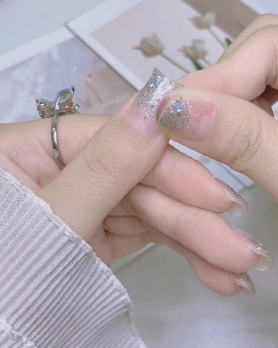 A hand folding the excess length of a sparkly silver nail sticker downward over the edge of the thumb nail.