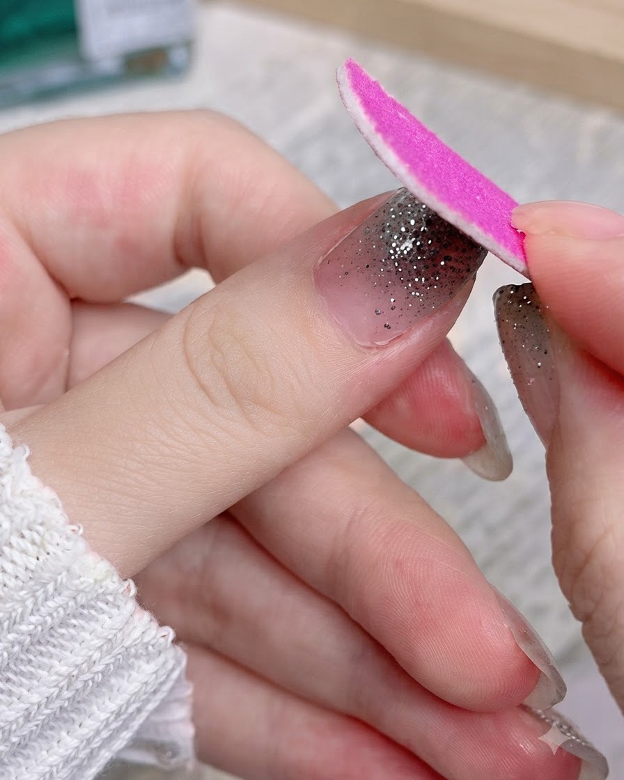 A pink emery board filing off the excess portion of a silver glitter nail sticker on a thumb.