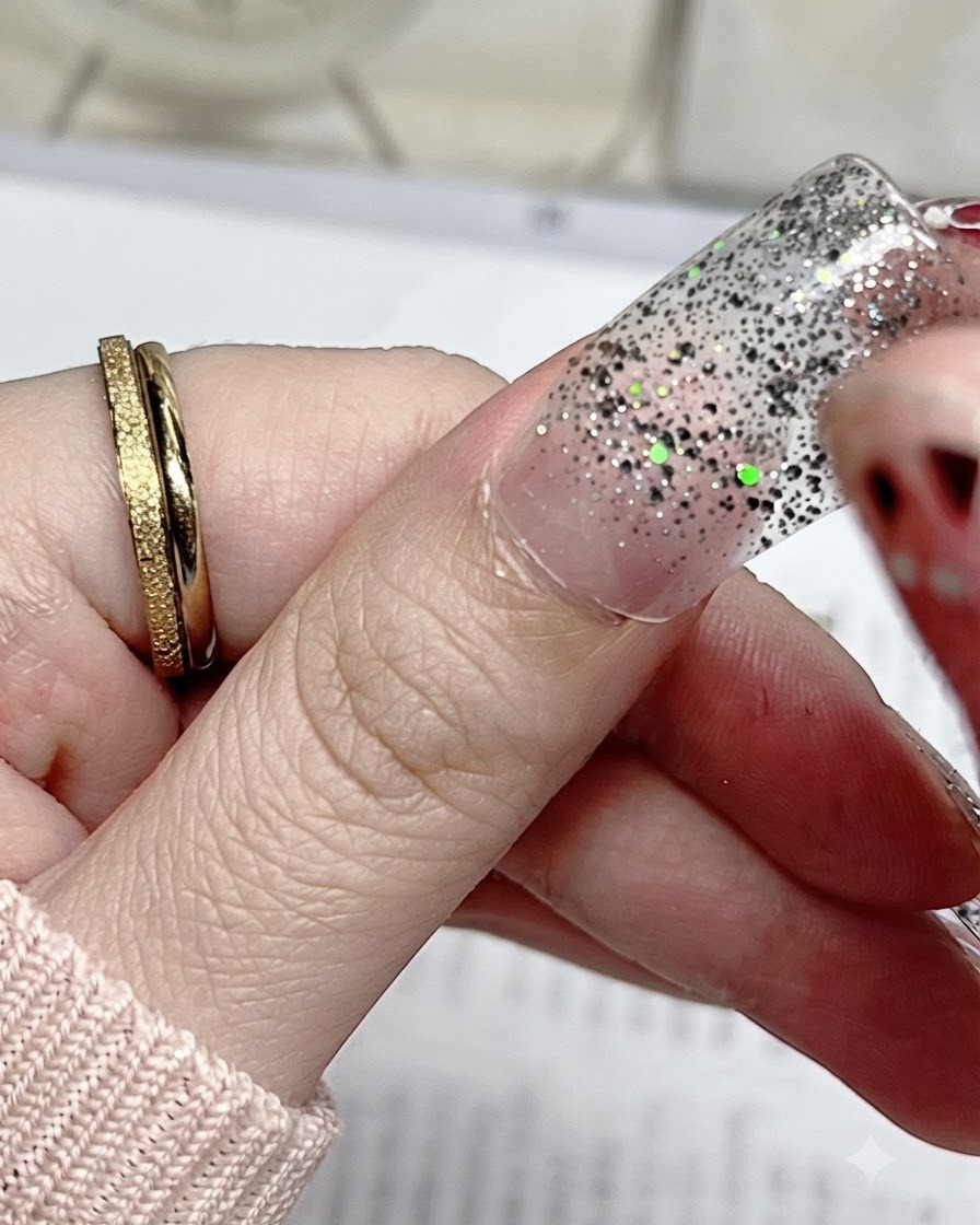 A close-up of a silver glitter nail sticker being aligned precisely with the cuticle of a natural thumb nail.