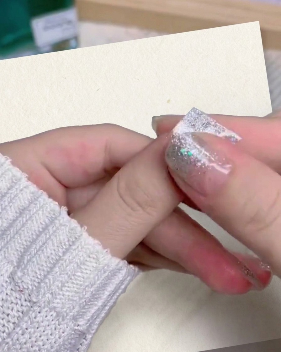 A close-up of a persons thumb and index finger firmly pressing and smoothing down a silver glitter gradient false nail onto their finger.
