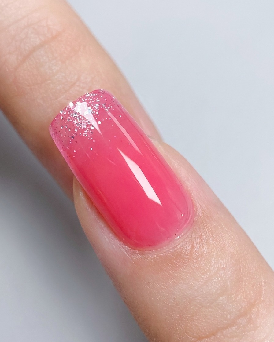 A close-up of a glossy pink gradient fingernail with a jar of silver glitter gel slightly blurred in the background.