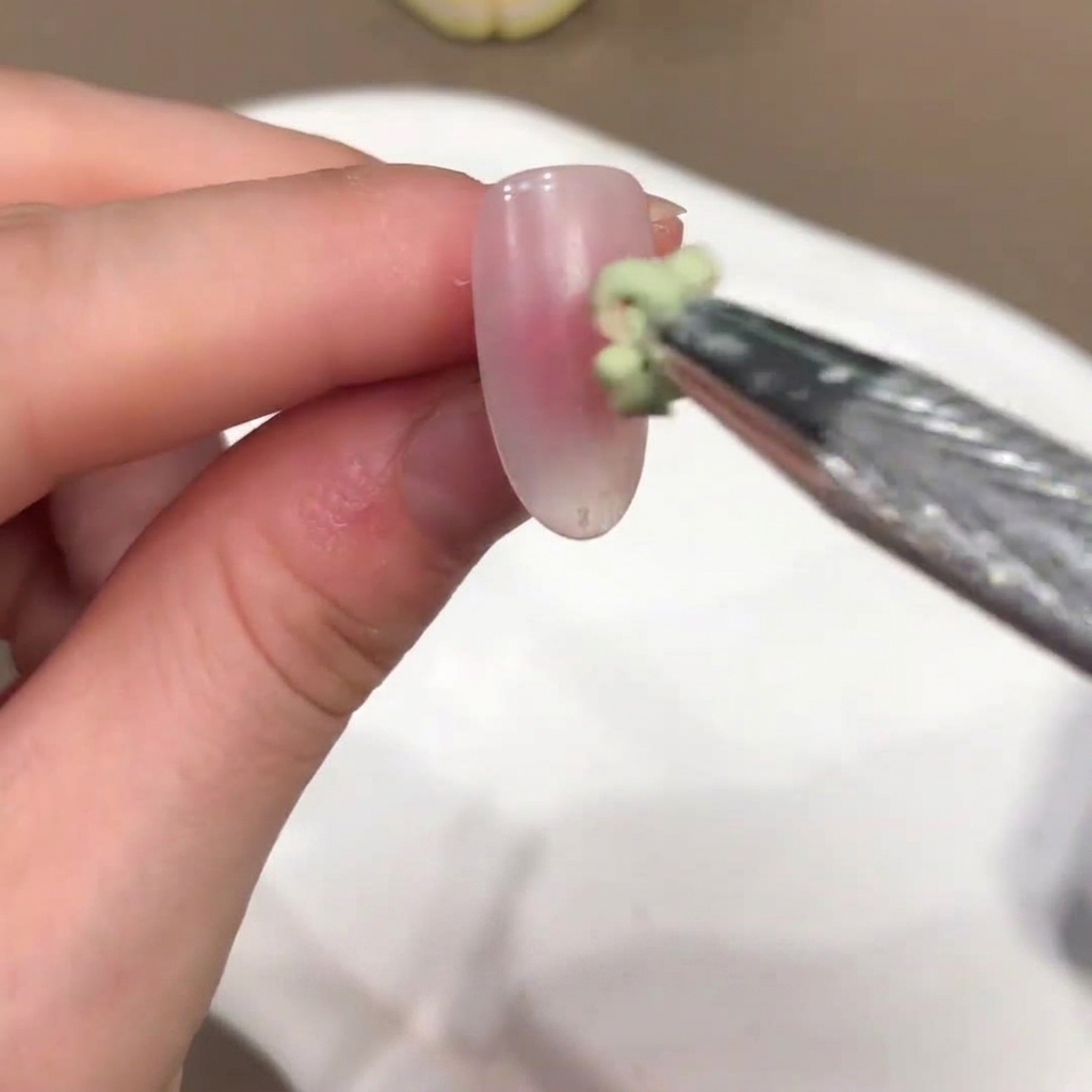 A small brush applying a green-tinted gel blob onto the center of a nude pink nail.