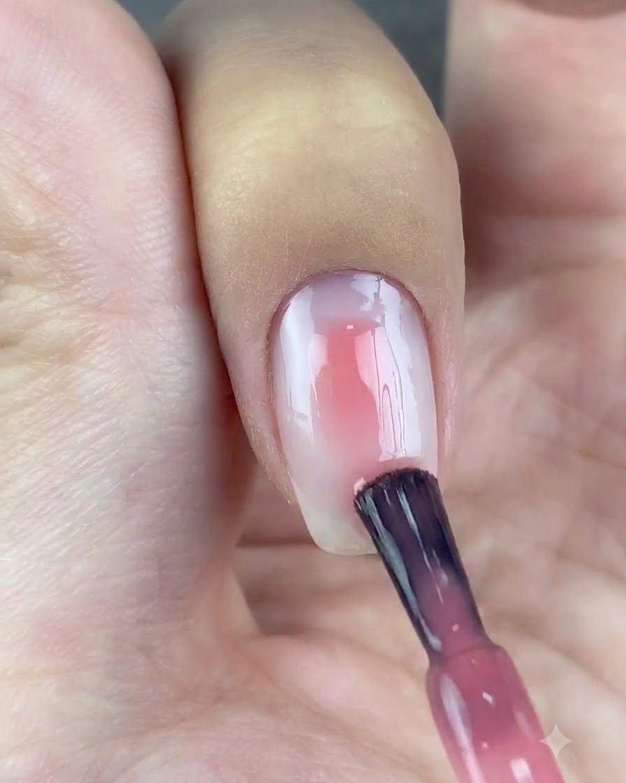 A polish brush applying a spot of translucent pink gel to the center of a white base nail.