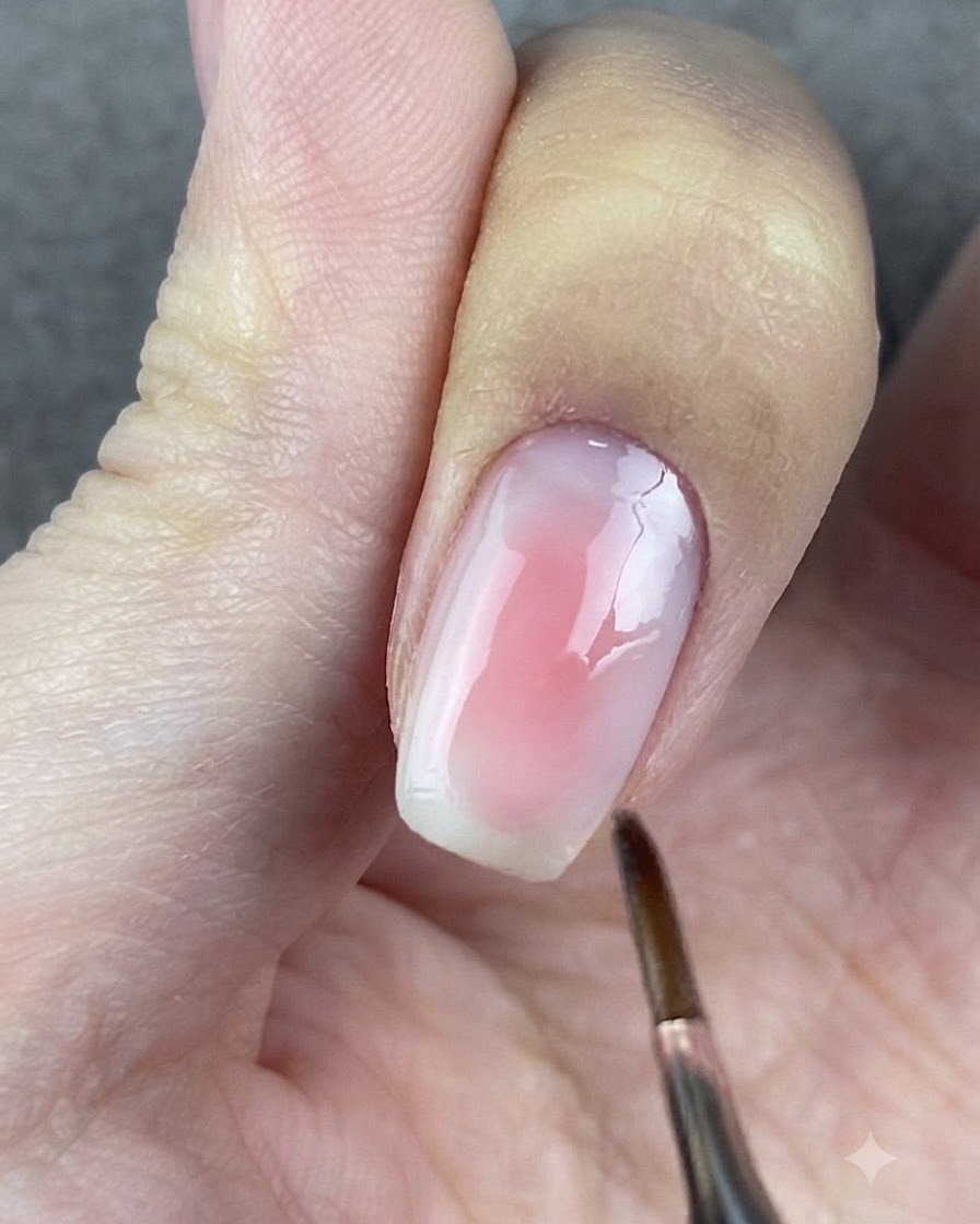 A round-headed brush gently tapping the edges of the pink gel on a fingernail.