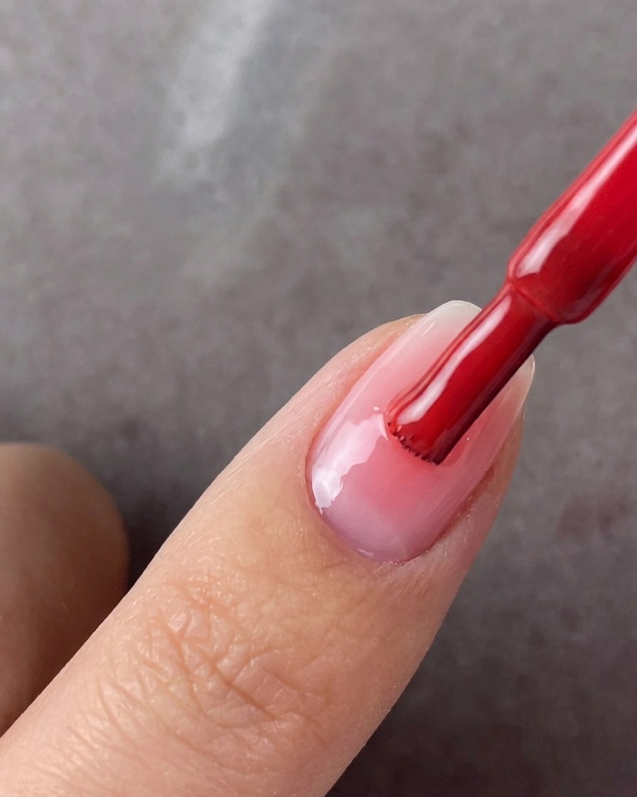 A red polish brush applying a second coat of pink gel to the center of a manicured nail.