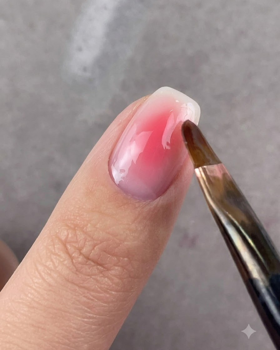 A round-headed brush gently blending the edges of pink gel on a fingernail.