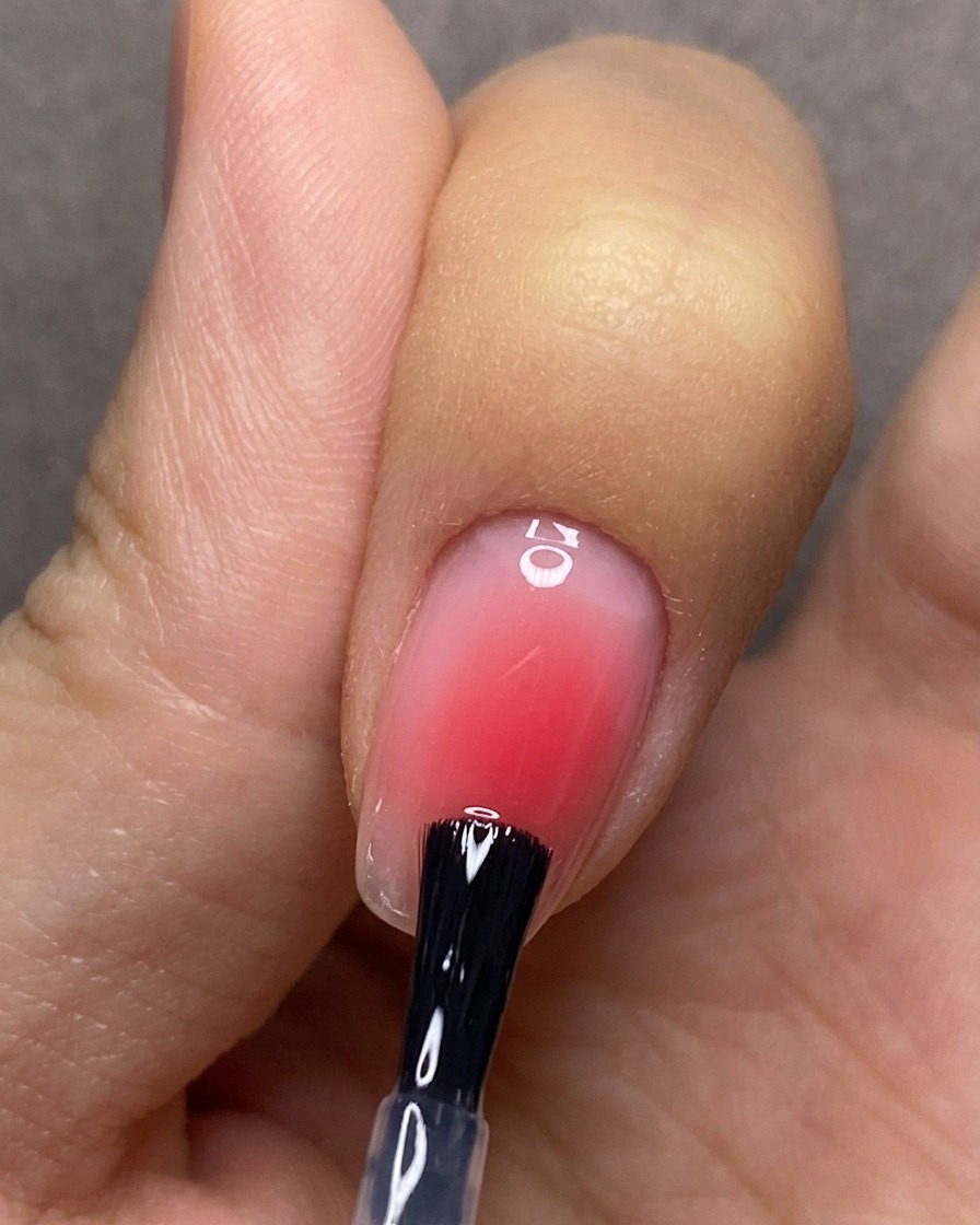 A brush applying a clear top coat evenly over a nail featuring a red blush gradient design.
