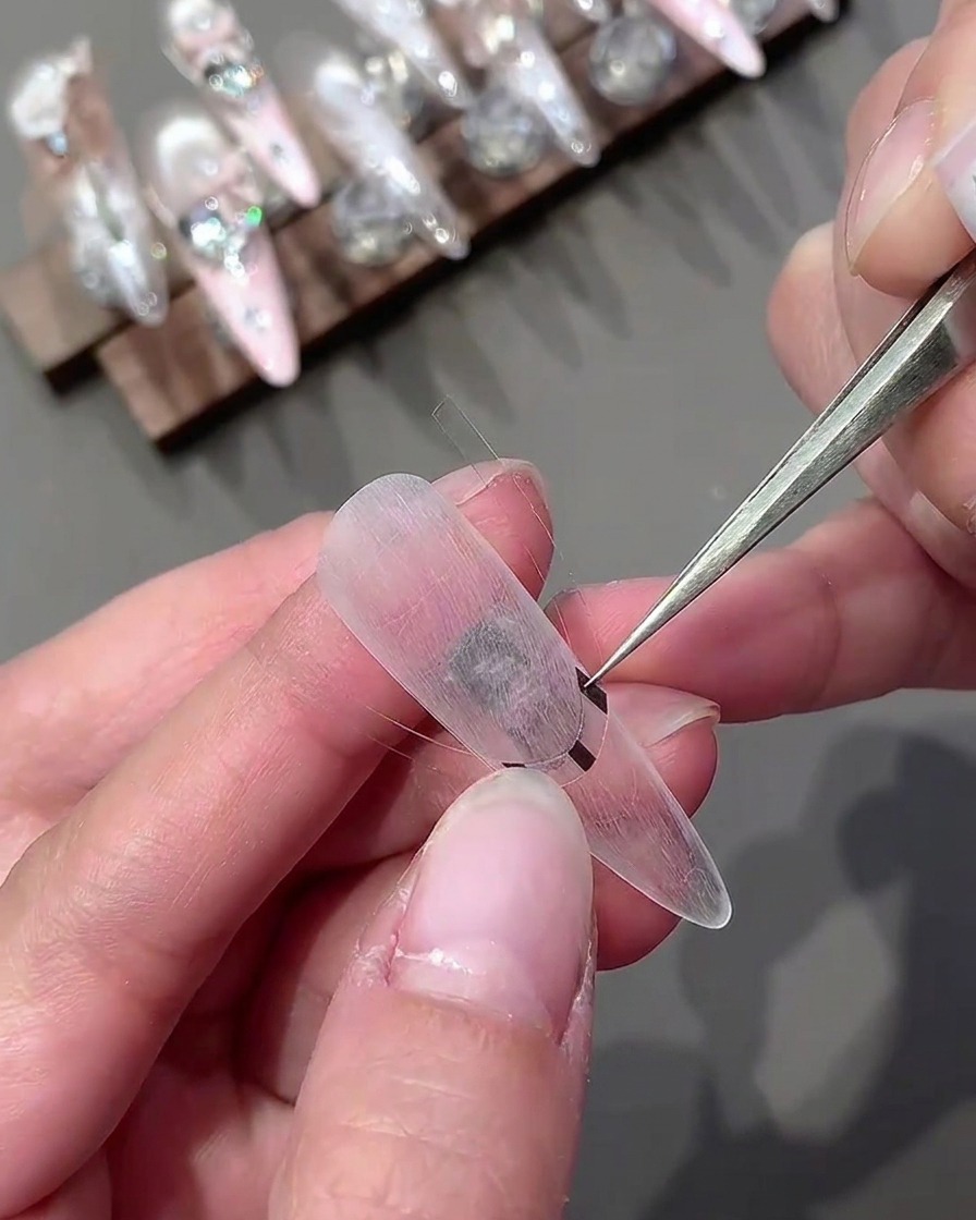 A pair of tweezers placing a curved sticker horizontally onto a clear nail tip.