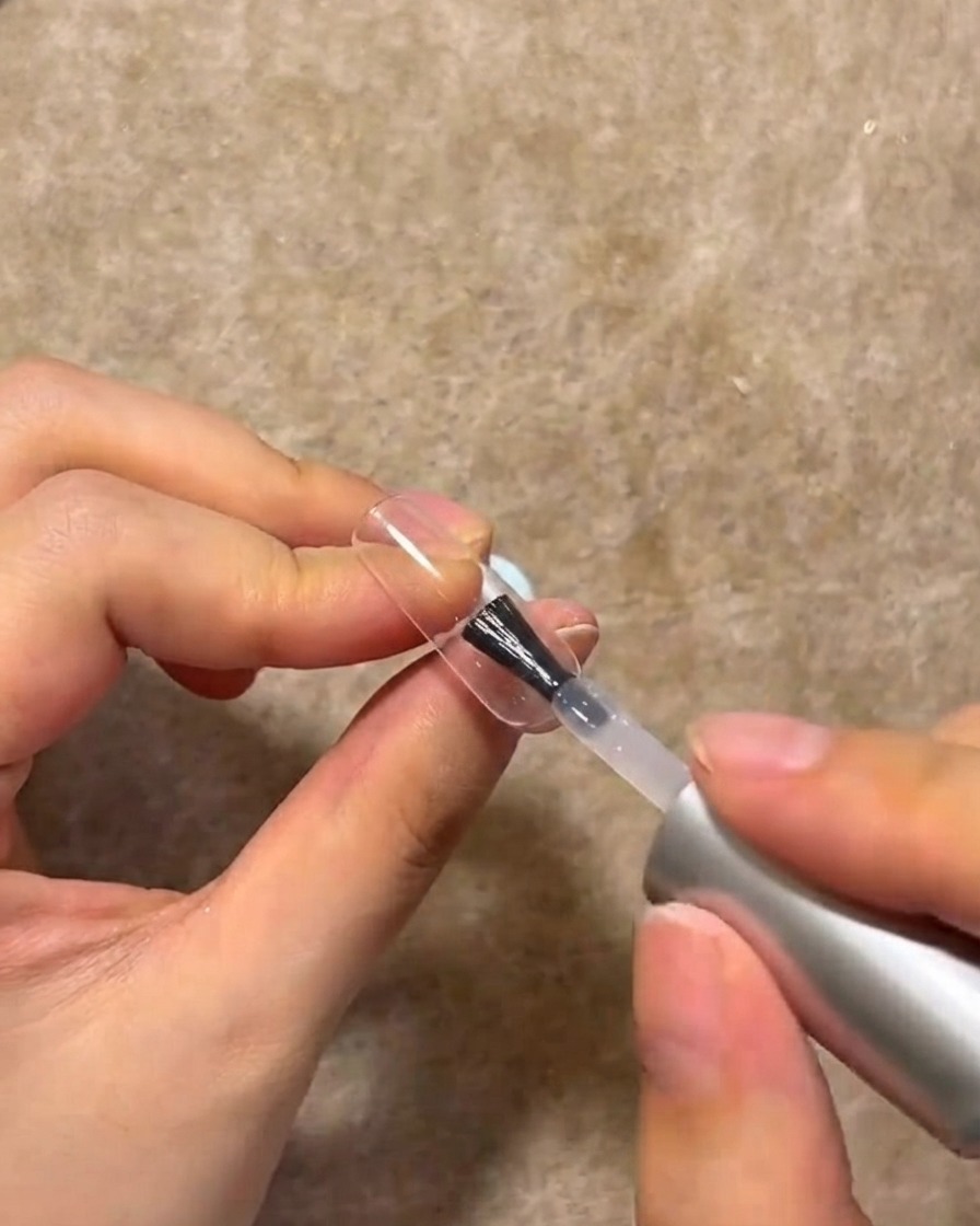 A nail brush applying a coat of clear building gel onto the surface of a manicured nail.