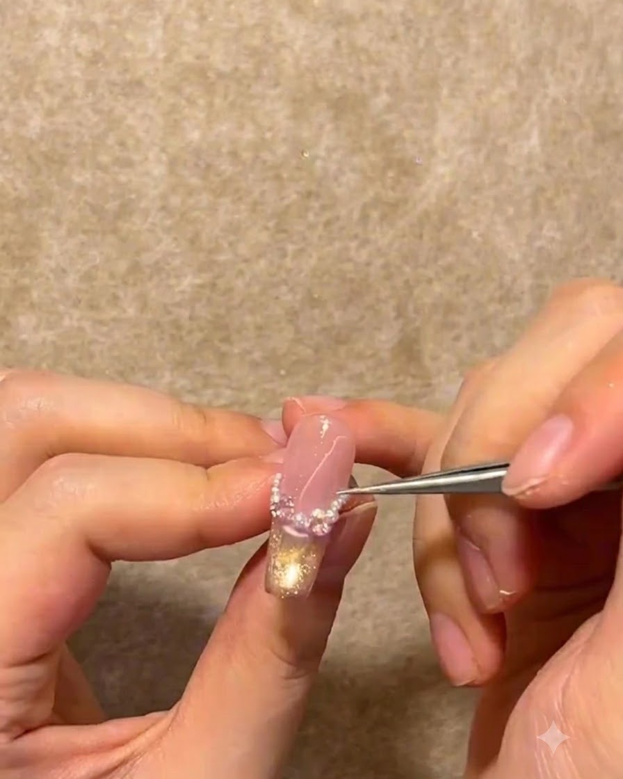 A pair of metal tweezers placing tiny white pearls and rhinestones along the French smile line of a nail.