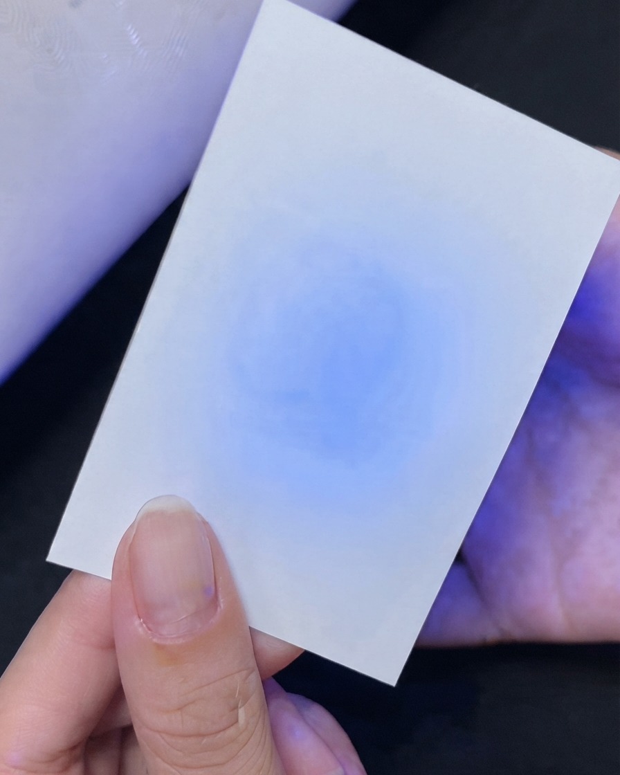 A hand holding a white card showing a blue UV light reflection test underneath.