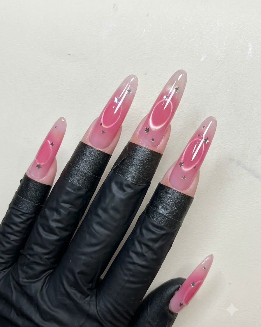 A nail brush applying a smooth layer of shiny top coat over long almond-shaped nails decorated with star sequins and cat eye art.