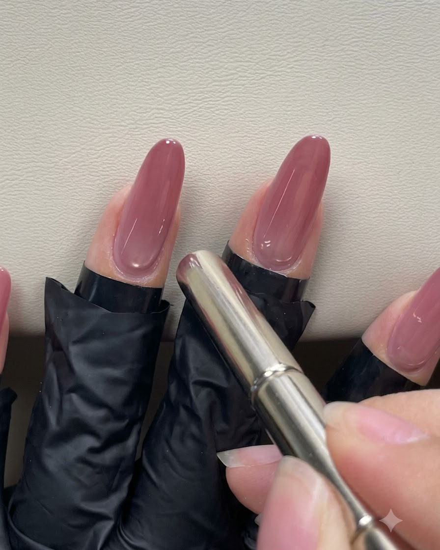 A hand using a cylindrical pen magnet, hovering it near the cuticle of a peach pink gel nail to manipulate the magnetic cat-eye highlight.