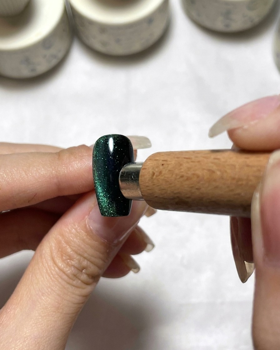 A magnetic tool positioned next to a dark green cat eye nail to distribute pigment.