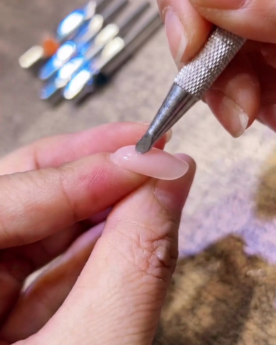 A close-up of a metal applicator tool applying pink blush pigment to the center of a nude gel nail.