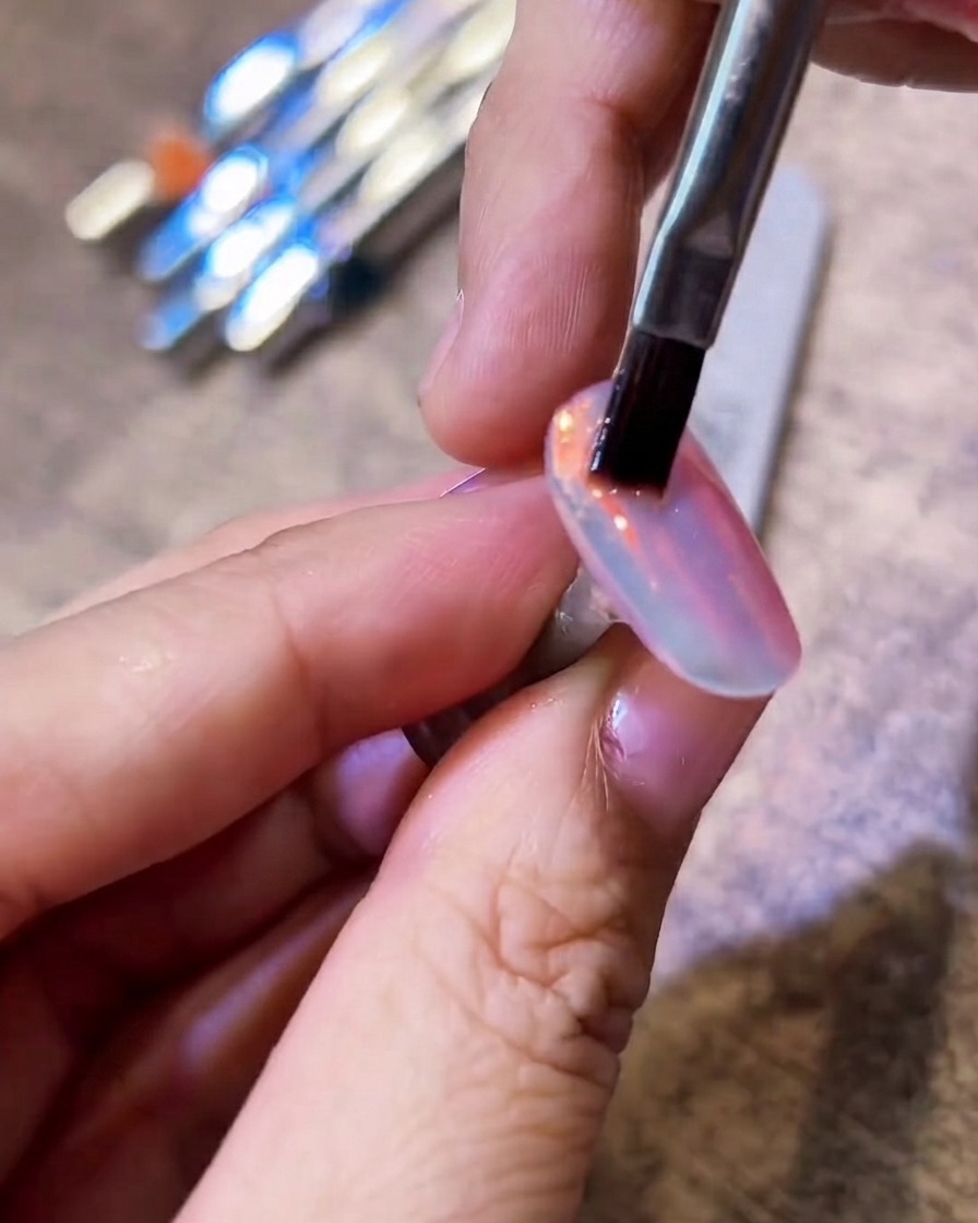 A flat bristle brush gently sweeping over a shiny aurora chrome nail to remove excess powder.