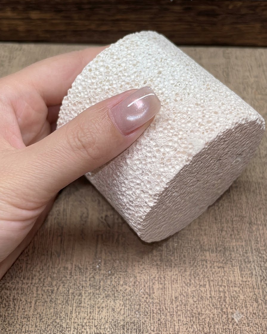 A close-up of a single nail resting against a textured white block, displaying a soft champagne glass bead cat-eye reflection.