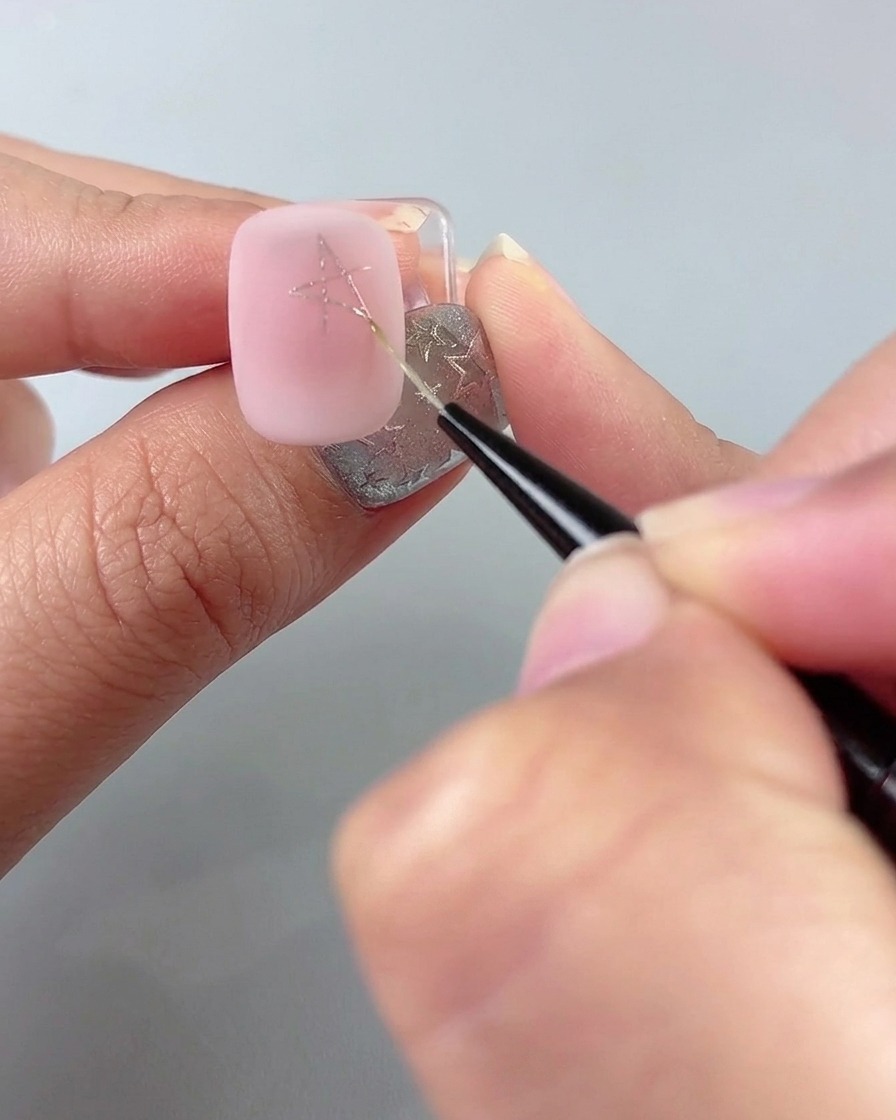 A fine liner brush drawing lines to complete a star shape on a pink manicured nail.