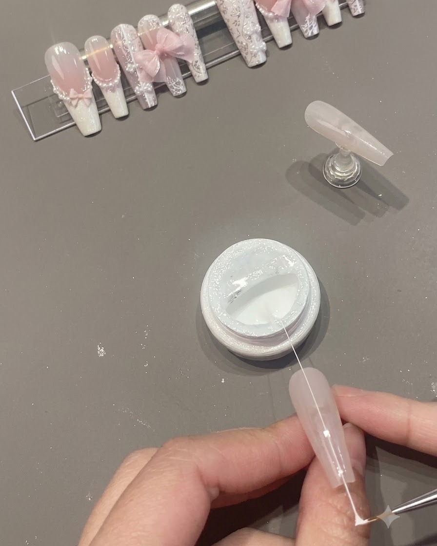 A narrow strand of white spider gel being pulled across the nail surface above an open jar.
