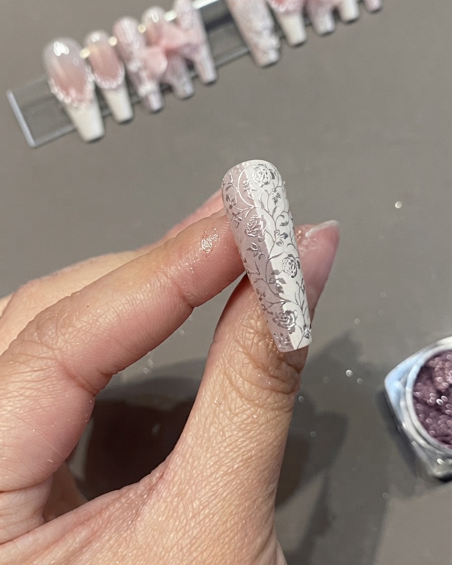 A close-up of a long coffin press-on nail featuring a detailed white floral lace and chrome pattern held between fingers.