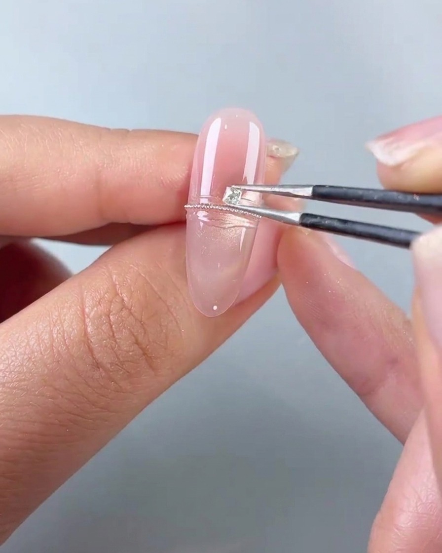 A pair of tweezers placing a small square diamond next to a line of steel beads on a pink gel nail.