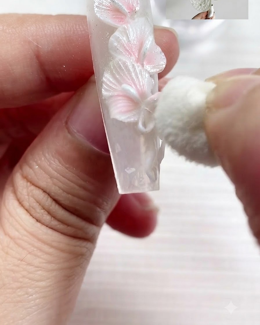 A small cosmetic sponge gently dabbing pearl white pigment onto the textured edges of sculpted white calla lilies and a bow on a nail.