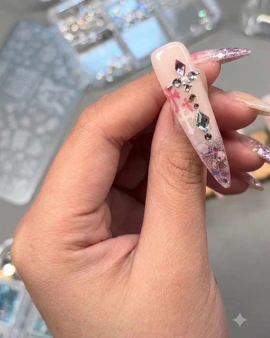 Close-up of a decorated nail adorned with crystals, a pearl, and metal charms over pink glitter and stamped art.