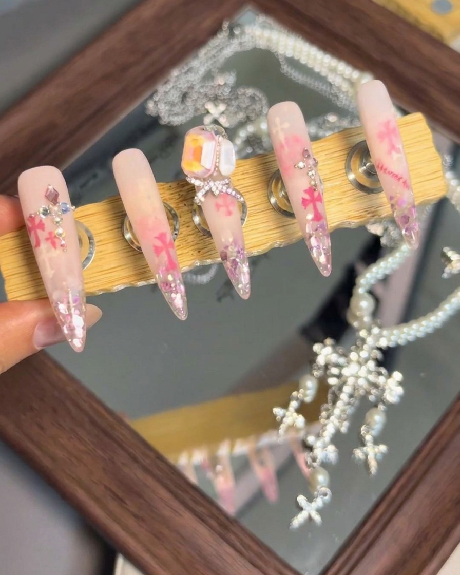 A display stand showcasing five finished press-on nails with elaborate crystal arrangements, bows, and Chrome Hearts accents.